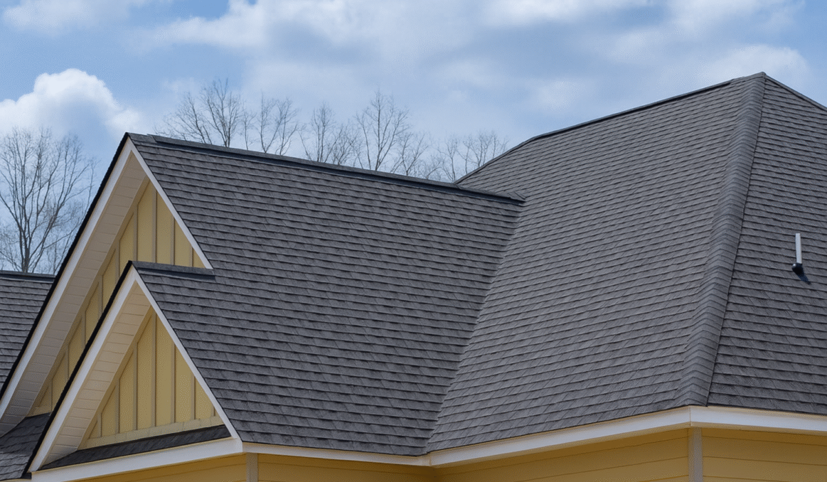 Close-up view of a residential home with dark gray asphalt shingle roofing, beige-yellow siding, and white trim, set against a blue sky with scattered clouds and bare trees in the background.