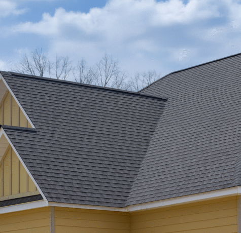 Close-up view of a residential home with dark gray asphalt shingle roofing, beige-yellow siding, and white trim, set against a blue sky with scattered clouds and bare trees in the background.