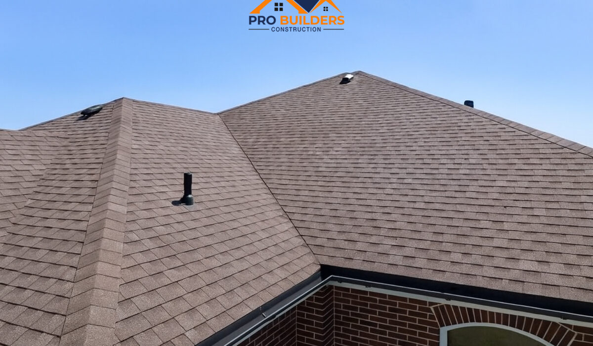 A brown shingle roof with multiple slopes and vent pipes on a brick house, under a clear blue sky.