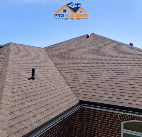 A brown shingle roof with multiple slopes and vent pipes on a brick house, under a clear blue sky.