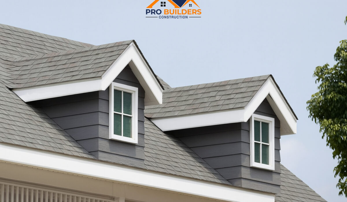 House roof with gray asphalt shingles and two dormer windows with white trim, set against a clear sky and partial tree in the background.