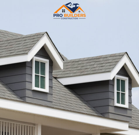 House roof with gray asphalt shingles and two dormer windows with white trim, set against a clear sky and partial tree in the background.