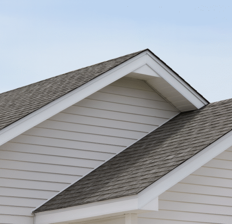 Close-up view of a modern house roof with gray asphalt shingles and white vinyl siding, shown from a slightly angled perspective under a clear blue sky.