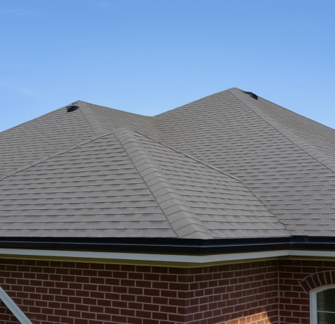 Gray asphalt shingle roof on a red brick house with minimal roof vents, viewed from an angled perspective under a clear blue sky.