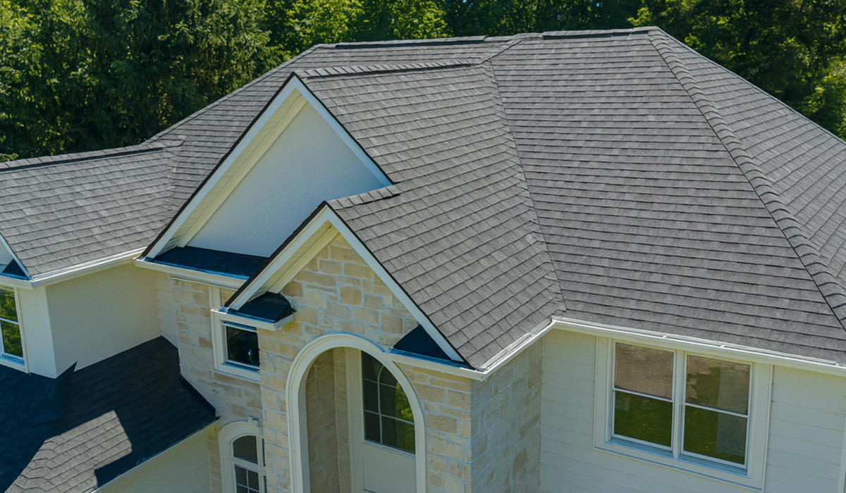 A high-resolution aerial view of a two-story house with light stone and white siding exterior, featuring a clean, dark gray shingle roof, surrounded by lush green trees.