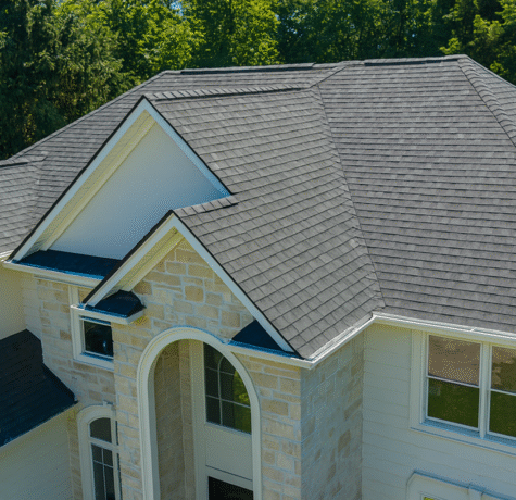 A high-resolution aerial view of a two-story house with light stone and white siding exterior, featuring a clean, dark gray shingle roof, surrounded by lush green trees.