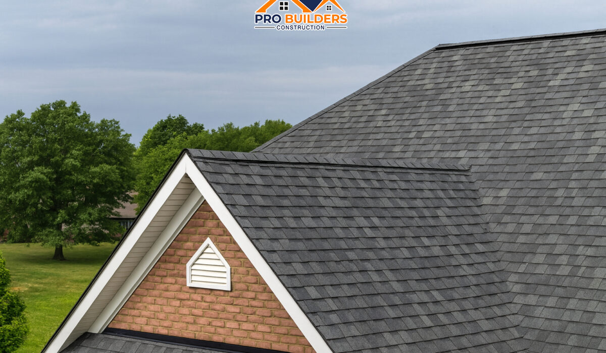 Brick gable end of a house with a gray asphalt shingle roof, featuring a small white gable vent at the peak, surrounded by green trees and a cloudy sky in the background.