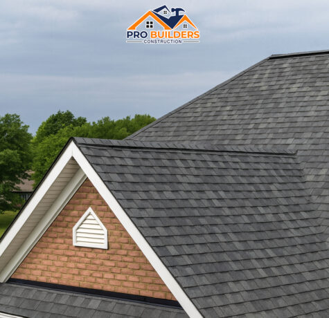 Brick gable end of a house with a gray asphalt shingle roof, featuring a small white gable vent at the peak, surrounded by green trees and a cloudy sky in the background.