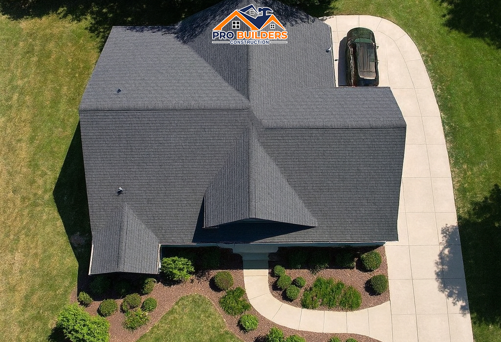 Aerial view of a modern single-story house with a dark gray shingle roof, surrounded by neatly trimmed shrubs, curved walkways, and a long concrete driveway leading to a parked car, set in a spacious green lawn.
