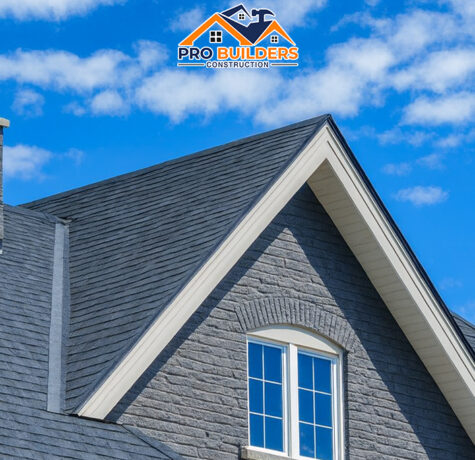 A contemporary home exterior featuring gray stone walls, pitched shingle rooflines, and white-trimmed windows against a partly cloudy sky.