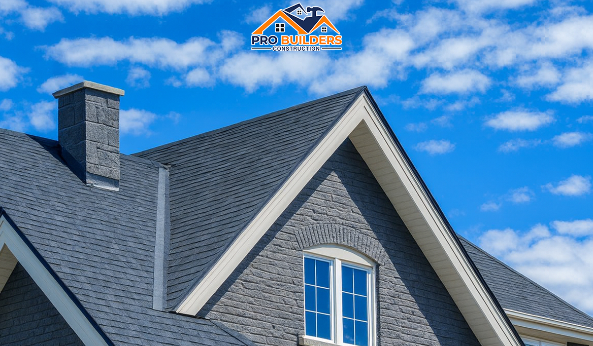 A contemporary home exterior featuring gray stone walls, pitched shingle rooflines, and white-trimmed windows against a partly cloudy sky.