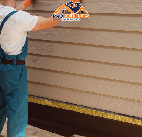 Construction worker in teal overalls and a yellow hard hat installing beige siding panels on a house exterior