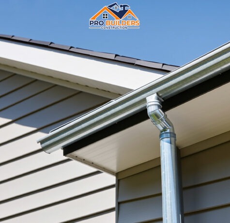 Close-up view of a home’s gutter system and downspout installed on vinyl siding, showing clean, well-fitted gutters under a clear blue sky - Pro Builders Construction.