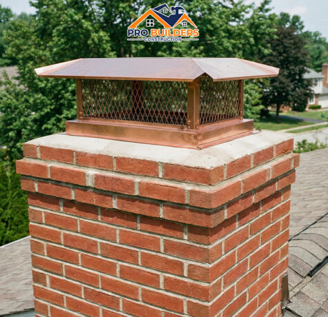 A close-up view of the top of a pristine red brick chimney on a residential roof. The chimney is topped with a shiny, copper-colored metal cap featuring a mesh spark arrestor. The background shows a sunny suburban neighborhood with green trees and the roofs of other American-style homes.
