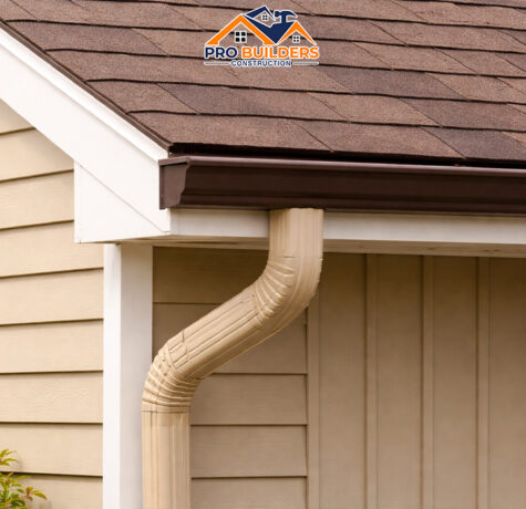 Close-up of a house roof corner with brown asphalt shingles, a dark bronze metal gutter, and a beige downspout attached to light tan horizontal siding under soft daylight.
