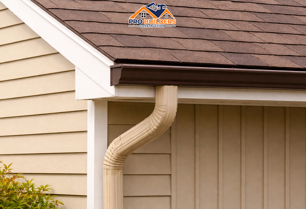 Close-up of a house roof corner with brown asphalt shingles, a dark bronze metal gutter, and a beige downspout attached to light tan horizontal siding under soft daylight.