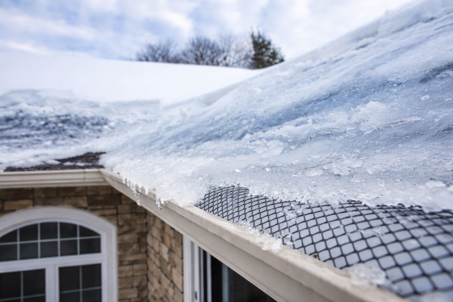 Ice buildup and snow covering a residential roof gutter with mesh guard, forming an ice dam along the roof edge in winter.